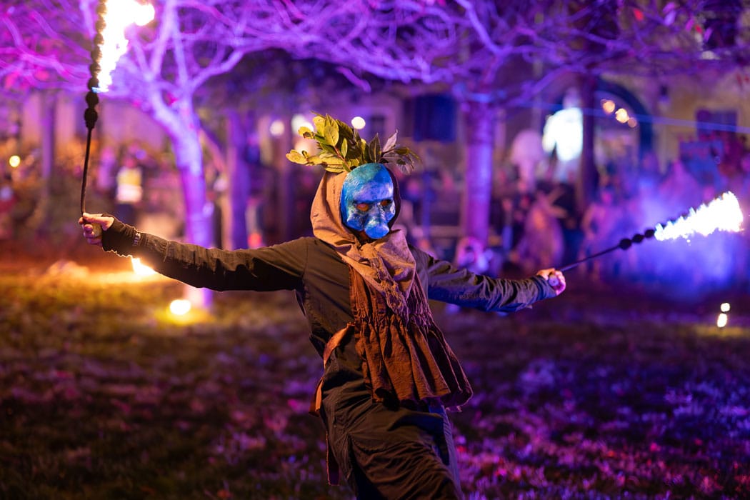 A street perfomer wearing a mask and swinging two fire sticks during a street carnival